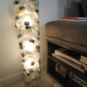 May include: A large, clear glass vase filled with frosted light bulbs, some lit, producing a warm light. The vase has a slight curve and is positioned on a tiled floor. The website address zerawfactory.sumupstore.com is displayed at the bottom.