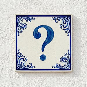May include: Square ceramic tile featuring a blue question mark on a white background. The tile has a blue border and decorative corner patterns. The question mark is centrally positioned.