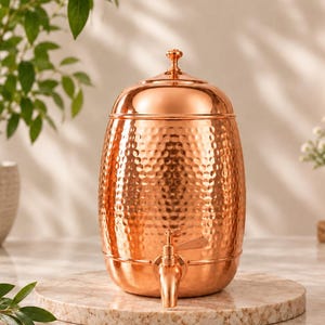 Puede incluir: Un dispensador de bebidas cilíndrico de cobre con textura martillada, acabado pulido y tapa a juego con un pequeño pomo. El dispensador incluye un grifo metálico funcional en la base para servir líquidos, mostrando un tono metálico cálido.