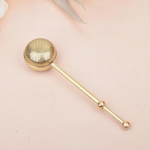 May include: A gold tea infuser with a perforated spherical basket and a long, slender handle. The infuser is designed for steeping loose leaf tea. The infuser is made of metal and has a polished finish. The handle has a decorative element.