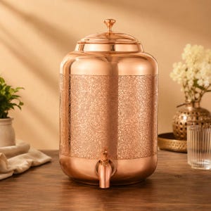 May include: A cylindrical copper water dispenser featuring a polished finish and an intricate, repeating floral pattern etched across the center. The vessel includes a matching domed lid with a small knob and a functional copper spigot at the base.