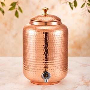 May include: A cylindrical copper water dispenser featuring a hammered texture across its surface. The vessel includes a removable lid with a small gold-toned knob handle and a silver-colored spigot at the base with the letters KK printed on it.