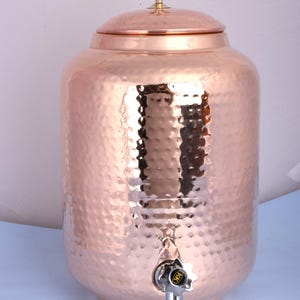 Puede incluir: Un dispensador de agua de cobre con textura martillada. El dispensador tiene una tapa con un pomo dorado y un grifo plateado. El cuerpo del dispensador es de color cobre claro. El dispensador está diseñado para contener agua.