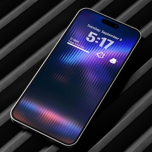 Puede incluir: Un smartphone con marco blanco muestra un fondo de pantalla abstracto y vibrante en tonos azules, morados y rosas. La pantalla muestra la hora 5:17, la fecha e iconos del tiempo. El teléfono descansa sobre una superficie negra acanalada.