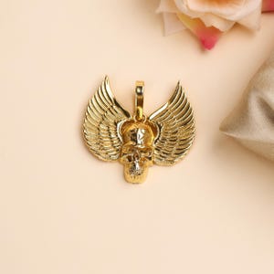 Peut inclure: Un pendentif doré représentant un crâne avec des ailes détaillées. Le pendentif a une boucle pour une chaîne. Les ailes sont finement conçues avec des détails de plumes. Le crâne a un visage détaillé.
