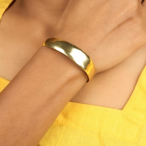 Puede incluir: Un brazalete de color dorado con una superficie lisa y ligeramente irregular. El brazalete se lleva en el brazo de una persona, y el fondo es una tela amarilla brillante.