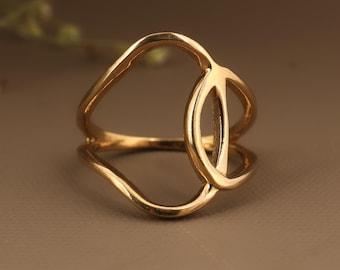 May include: A gold-colored ring with an open, geometric design. The ring features a double band with an abstract shape in the center. The ring is made of a shiny, metallic material and is set against a neutral background.