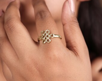 May include: A gold ring with an elaborate knot design. The ring is worn on a finger, highlighting its detailed craftsmanship and elegant appearance. The knot design is the focal point, adding a touch of sophistication to the jewellery.