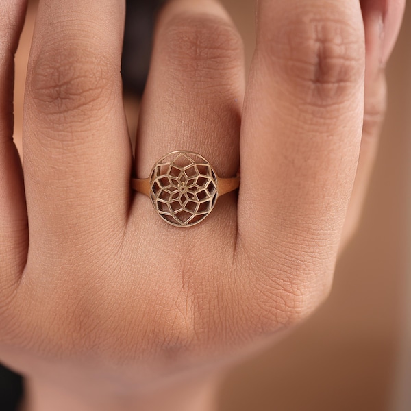 Peut inclure: Une bague dorée avec un motif géométrique ajouré. La bague présente un élément central circulaire avec un motif symétrique détaillé. La bague est portée au doigt, mettant en valeur le savoir-faire de la bijouterie.