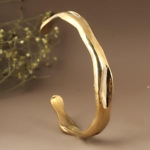 May include: A gold-coloured, open cuff bracelet with an organic, flowing design. The metal has a textured surface and a slightly irregular shape. The bracelet is a statement piece of jewellery, with a smooth, polished finish.