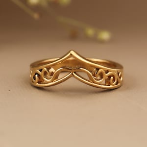 May include: A gold ring with a distinctive curved, v-shaped design. The band is adorned with detailed scrollwork, giving the ring an elegant and ornate look. The ring is likely made of gold or a gold-toned metal.