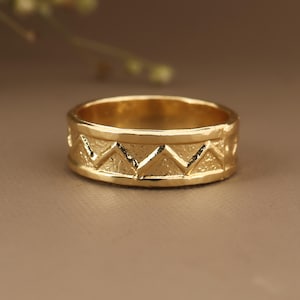 Puede incluir: Un anillo dorado con una superficie texturizada y un patrón geométrico repetitivo de triángulos invertidos. El anillo es de estilo banda, con una anchura de aproximadamente 0,6 cm. El anillo se muestra sobre un fondo neutro.