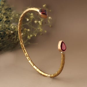May include: A gold-coloured cuff bracelet with a hammered texture. The bracelet features two teardrop-shaped, deep red gemstones set in gold bezels. The open design allows for easy wear and adds a touch of elegance.