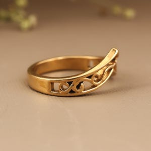 May include: A gold-colored ring with an openwork design. The ring features a curved band with a decorative, swirling pattern. The ring's design includes cut-out shapes and a raised section on one side. The ring is likely made of metal.