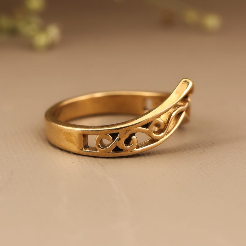 May include: A gold-colored ring with an openwork design. The ring features a curved band with a decorative, swirling pattern. The ring's design includes cut-out shapes and a raised section on one side. The ring is likely made of metal.