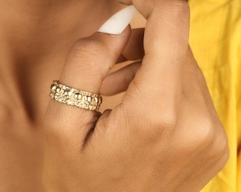 May include: A gold-colored ring with a textured design featuring small, raised circular elements. The ring is worn on a finger, with a yellow garment visible in the background. The ring's design is intricate and detailed.