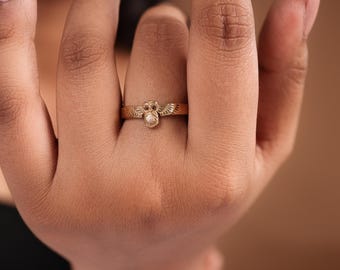 May include: A gold-colored ring featuring an owl design. The ring has a detailed owl figure at the center, with wings extending on either side. The ring is worn on a finger, showcasing the intricate details of the jewelry.