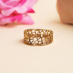 May include: A gold-colored ring with a geometric design of interconnected triangles. The ring is wide and has an open, airy appearance. The background features a blurred pink flower and a soft, neutral surface.