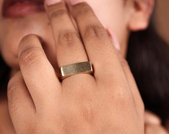 May include: A gold-colored rectangular ring. The ring is a simple, minimalist design with a flat top and sides. The ring is worn on a finger.
