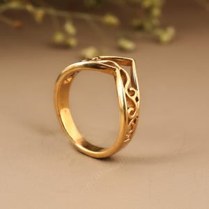 May include: A gold-colored ring with an open, geometric design. The ring features intricate, swirling filigree patterns along the side. The ring is set against a neutral background, highlighting its polished finish. This piece is a jewelry item.