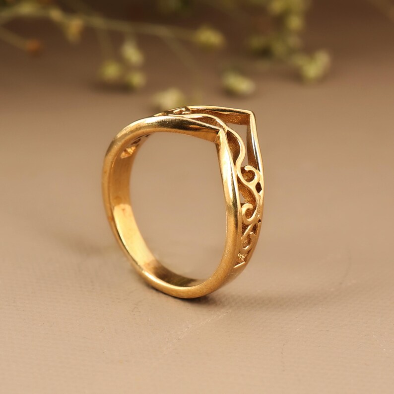 May include: A gold-colored ring with an open, geometric design. The ring features intricate, swirling filigree patterns along the side. The ring is set against a neutral background, highlighting its polished finish. This piece is a jewelry item.