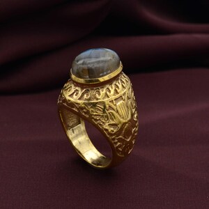 May include: A gold ring with a grey oval gemstone. The ring has detailed embossed floral and scrollwork patterns. The gemstone is set in a gold bezel, and the ring has a wide band.
