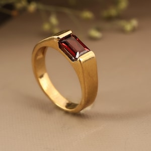 Könnte beinhalten: Ein goldfarbener Ring mit einem rechteckigen, tiefroten Edelstein. Der Ring hat ein schlichtes, elegantes Design, wobei der Edelstein in einer Zargenfassung gefasst ist. Das Band ist glatt und poliert.