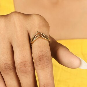 May include: A gold-colored ring with a V-shaped design. The ring features an openwork pattern with decorative swirls along the band. The ring is worn on a finger against a yellow background.
