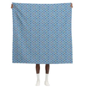 May include: A light blue scarf with a repeating pattern of small, colorful shapes. The shapes include blue, orange, yellow, and green elements. The scarf is held up by a person.