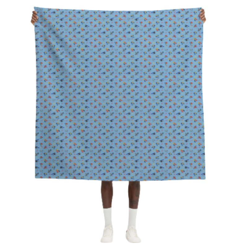 May include: A light blue scarf with a repeating pattern of small, colorful shapes. The shapes include blue, orange, yellow, and green elements. The scarf is held up by a person.