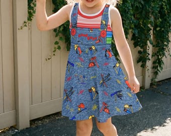 May include: A blue denim-style overall dress featuring a colorful pattern of construction tools and the text "good guys" on the chest pocket. The dress is worn over a sleeveless shirt with horizontal red, white, and green stripes.