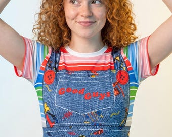 May include: A blue denim-print overall dress featuring red buttons and the text Good Guys on the chest pocket. The dress is decorated with a colorful pattern of tools and cowboy boots. It is worn over a short-sleeved shirt with horizontal stripes.