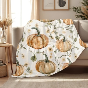 Peut inclure: Une couverture blanche avec un motif aquarelle de citrouilles, de vignes et de petites fleurs jaunes. Les citrouilles sont orange et marron avec des tiges vertes. La couverture est drapée sur un canapé beige.