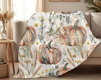 May include: A white throw blanket featuring a repeating pattern of watercolour pumpkins, wheat, and wildflowers in orange, green, and yellow hues. The blanket is draped over a beige sofa, creating a cosy autumn theme.