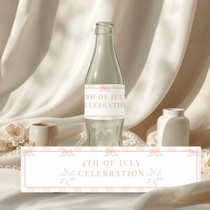 May include: A clear glass bottle with a label that reads "4th of July Celebration." The label has a pink and white striped pattern with bow accents. The bottle is surrounded by decorative elements, including a small vase and pearls.