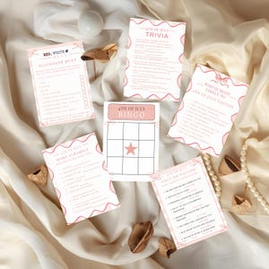 May include: A collection of 4th of July themed party games, including trivia, bingo, and a scavenger hunt. The cards have a white background with pink decorative borders and text. A pearl necklace and wooden decorations are also present.