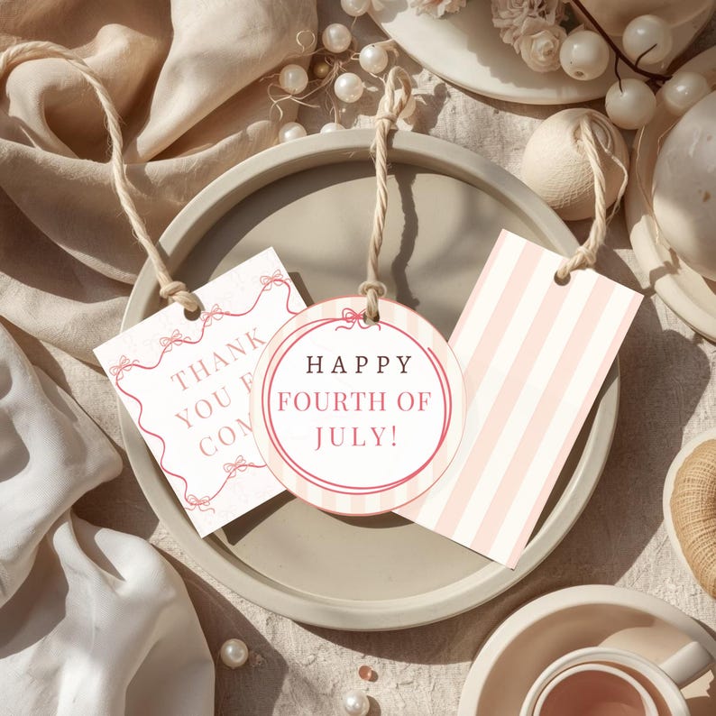 May include: Three gift tags with twine strings rest on a gray plate. One tag says "Thank You" with bows, another says "Happy Fourth of July!" with pink stripes, and the third has pink stripes. The tags are light pink and white.