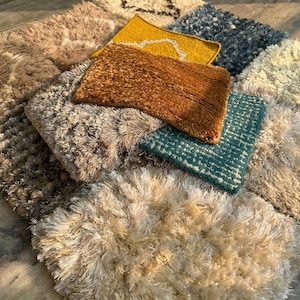 Könnte beinhalten: Eine Sammlung kleiner, strukturierter Teppiche in verschiedenen Farben, darunter Beige, Braun, Gelb und Türkis. Die Teppiche haben einen zotteligen oder geschlungenen Flor, der ein weiches und einladendes Aussehen erzeugt. Die Anordnung deutet auf eine Ausstellung von Wohnaccessoires hin.