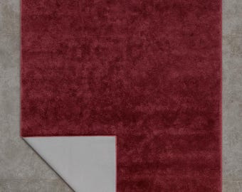 Puede incluir: Una alfombra rectangular de color burdeos con una superficie afelpada y texturizada. La esquina de la alfombra está doblada, revelando un reverso gris claro. La alfombra está colocada sobre un suelo de baldosas grises.