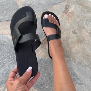 Pode incluir: Sandálias de couro preto com presilha para o dedo do pé e tira no tornozelo. As sandálias rasas têm um design minimalista. A imagem mostra um par de sandálias, uma sendo segurada e a outra usada no pé.