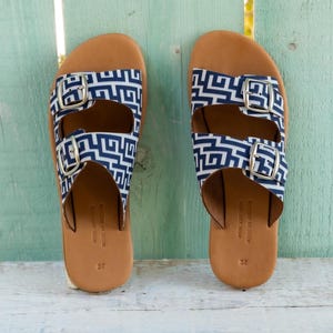 Puede incluir: Un par de sandalias con plantillas de cuero marrón y correas con estampado geométrico azul marino y blanco. Las correas tienen hebillas plateadas. Las sandalias se exhiben sobre una superficie de madera blanca sobre un fondo verde claro.