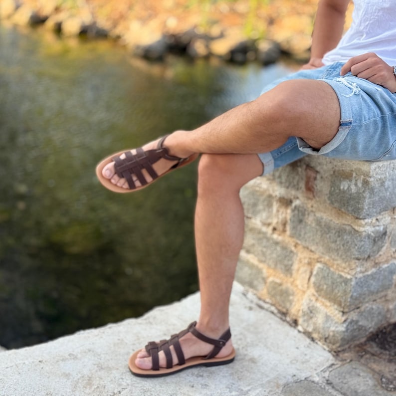 Pu&ograve; includere: Sandali in pelle marrone con design intrecciato. I sandali hanno una suola marrone chiaro e un cinturino alla caviglia. I sandali sono indossati da una persona seduta su un muro di pietra vicino all'acqua.