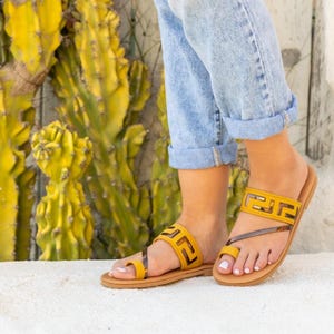 Puede incluir: Sandalias de cuero amarillas con un diseño geométrico en las correas. Las sandalias tienen una suela marrón y una correa para el dedo gordo. El fondo presenta un cactus amarillo y una pared blanca. La persona lleva vaqueros azules.