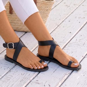 Peut inclure: Une paire de sandales plates noires avec un design à boucle d'orteil, une large bride de cheville dotée d'une boucle en métal argenté et une semelle simple. Les sandales sont portées avec un pantalon blanc sur une surface en bois clair.