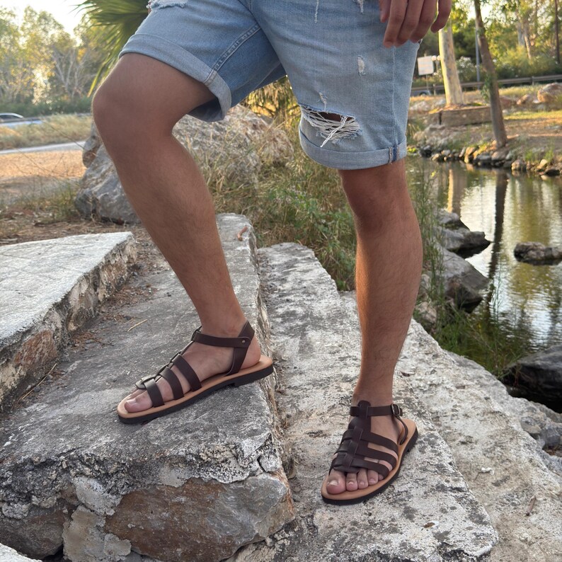 Pu&ograve; includere: Sandali in pelle marrone con pi&ugrave; cinturini e chiusura con fibbia. I sandali hanno una suola marrone chiaro e sono indossati con pantaloncini di jeans blu chiaro. I sandali sono mostrati su una scala di pietra.