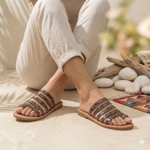 May include: Tan leather sandals with a snakeskin pattern on the straps. The sandals have a flat sole and a toe loop design. The person is wearing light-coloured trousers. The sandals are a comfortable, casual footwear option.