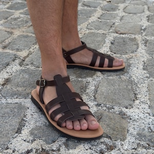 Pu&ograve; includere: Sandali in pelle marrone con design intrecciato e cinturini alla caviglia. I sandali hanno una suola marrone chiaro e una chiusura con fibbia. L'immagine mostra i sandali su una superficie di ciottoli.