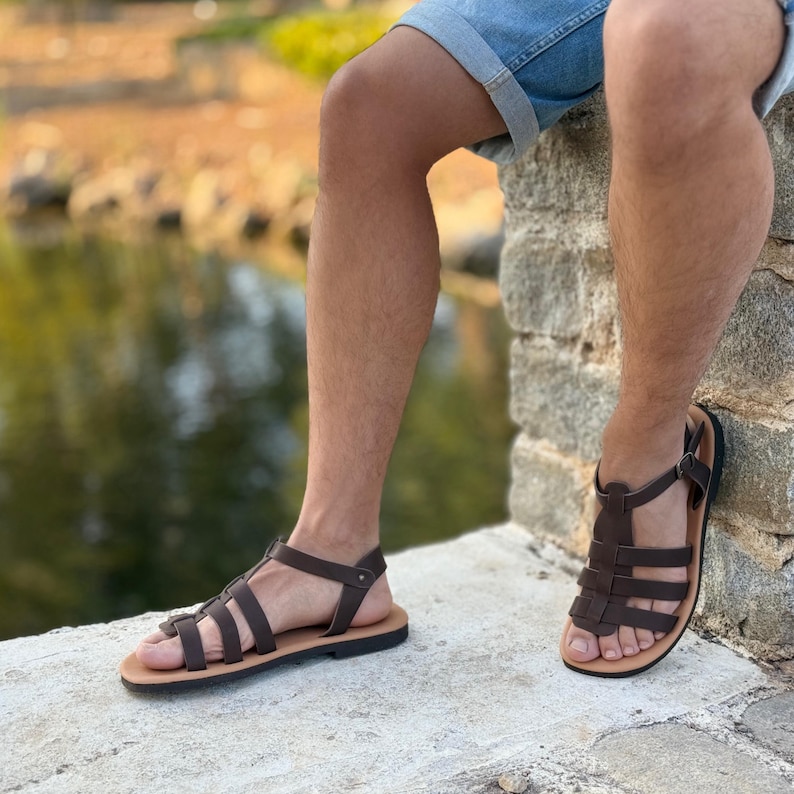 Pu&ograve; includere: Sandali in pelle marrone con pi&ugrave; cinturini e chiusura con fibbia. I sandali hanno una suola marrone chiaro e una tomaia marrone scuro. I sandali sono indossati da una persona seduta su un muro di pietra.