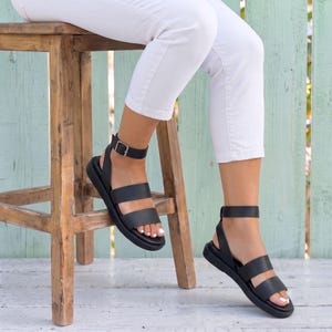 May include: A pair of black sandals featuring two horizontal straps across the foot and an adjustable ankle strap with a metal buckle. The sandals have a thick, flat sole and are worn with white pants, showcasing a simple and functional design.