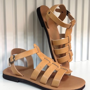 Pu&ograve; includere: Un paio di sandali in pelle color cuoio con pi&ugrave; cinturini e chiusura con fibbia. I sandali hanno una suola marrone scuro e sono esposti su una superficie bianca. Il design ricorda le calzature romane antiche.